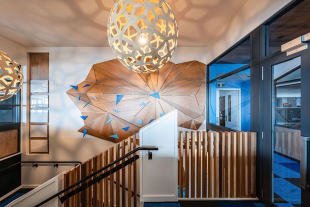 A contemporary stairway and landing with a dramatic geometric timber feature wall, blue angular accents, vertical timber balustrades, black metal handrails, and large perforated sculptural pendant lights. Glazed doors and windows open to adjacent spaces, with strong blue and warm wood tones creating a bold, artistic interior.