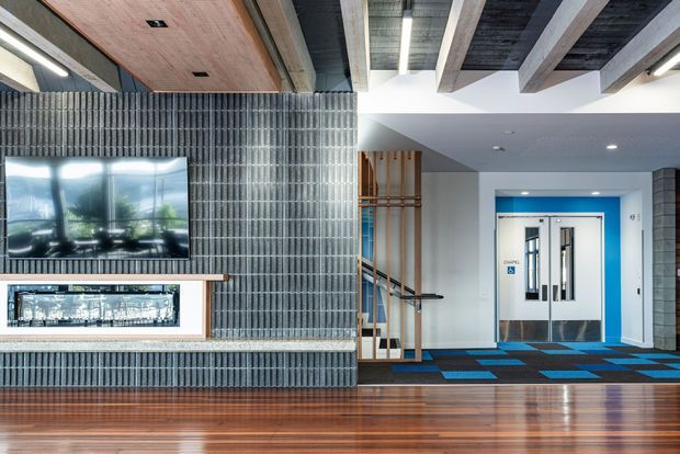 A contemporary commercial interior lobby or reception area with a dramatic exposed-beam ceiling, polished timber flooring, and a feature wall clad in vertically arranged dark grey tiles or slats. A wall-mounted television and illuminated display niche sit above a built-in bench or counter. The space includes a glazed timber screen, blue-accented elevator or doorway surrounds, and geometric blue and charcoal carpet tiles.