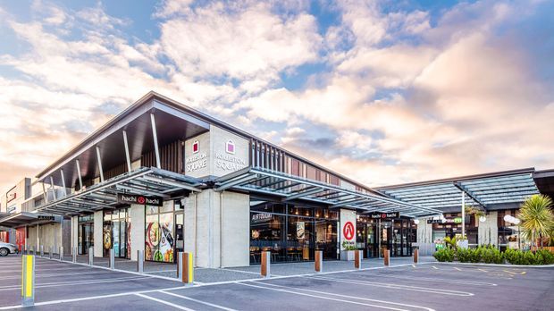 Exterior view of a contemporary low-rise retail and hospitality complex with covered pedestrian canopies, exposed steel framing, extensive glazing, and multiple shopfronts including restaurants and retail tenants. The building sits beside a marked parking area and is photographed under a partly cloudy sky.