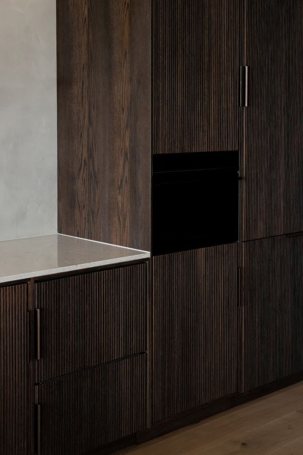 Close-up of a dark timber kitchen or built-in joinery installation featuring vertically fluted cabinet fronts, integrated dark handles, a black open appliance or storage niche, a pale stone benchtop, and a smooth light grey wall. Timber flooring is visible at the base.