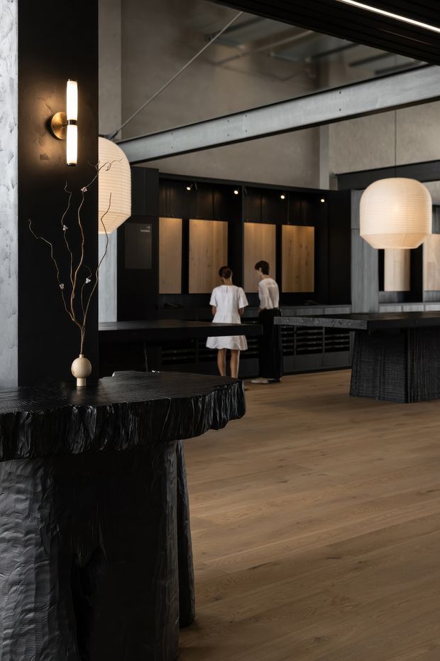 A dramatic, dark-toned hospitality or commercial interior with a bar and open kitchen area. The space features black textured counters and joinery, warm timber cabinet fronts, exposed concrete walls and ceiling structure, black metal framing, timber flooring, and oversized ribbed pendant lights. A sculptural black reception or bar table with a decorative branch arrangement occupies the foreground, while staff stand behind the central counter.
