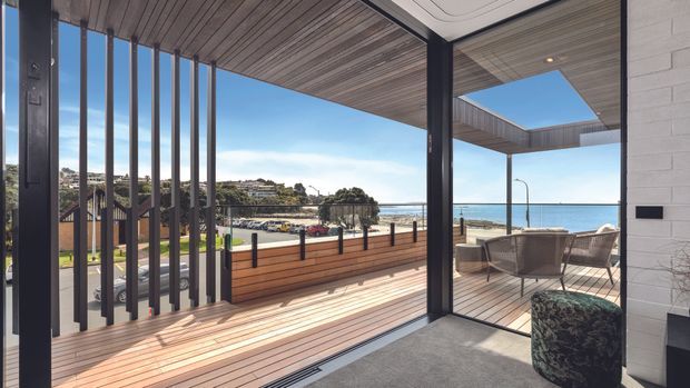A contemporary coastal terrace viewed from inside through large glazed openings. The outdoor deck features warm timber decking, a timber privacy planter wall, dark vertical screening, outdoor seating, and a pergola with a rectangular skylight. A bright ocean horizon and coastal streetscape are visible beyond.