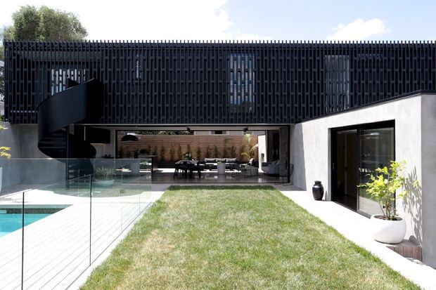 Contemporary courtyard house viewed from the garden, with a strong black vertical-screen upper facade, white rendered walls, extensive glazing, and a sculptural black spiral stair. A glass pool fence borders a turquoise swimming pool and pale deck, while the covered ground-floor living and dining area opens directly onto a central lawn courtyard.