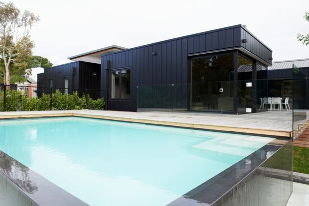A modern single-storey house with dark vertical metal-clad walls, extensive floor-to-ceiling glazing, and a flat roof surrounding a bright turquoise swimming pool. A paved pool terrace, glass pool fencing, outdoor dining furniture, and landscaped planting are visible. The image is taken from the outdoor pool area, with the building facade as the backdrop.