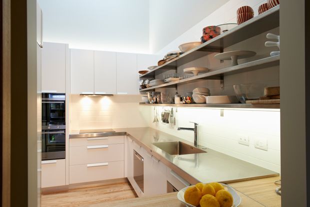 A bright, contemporary galley-style kitchen viewed from the entrance. White full-height cabinetry, integrated appliances, pale benchtops, a stainless-steel double sink, and a white tiled splashback create a clean, streamlined fit-out. Open timber shelves display crockery and cookware, while warm under-cabinet lighting and timber flooring add warmth.