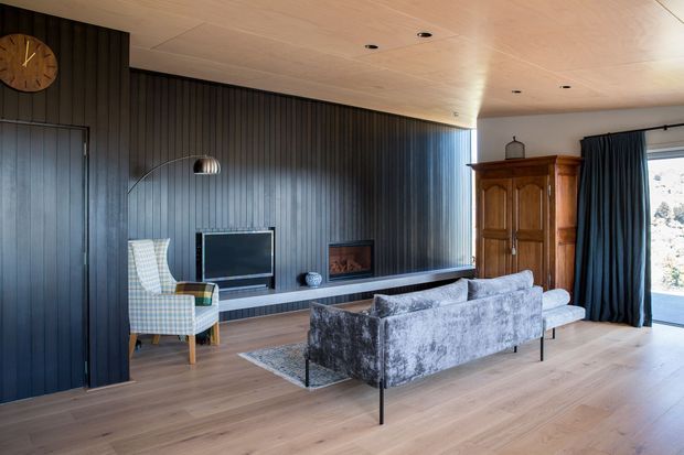 A refined living room with dark vertical timber wall panelling, a long built-in media and fireplace wall, grey velvet sofas, a patterned armchair, and a large traditional timber armoire. The room has pale timber flooring and a matching timber-lined ceiling, with dark curtains framing generous glazing and natural light. A minimalist arched floor lamp and wall clock complete the warm, sophisticated interior.
