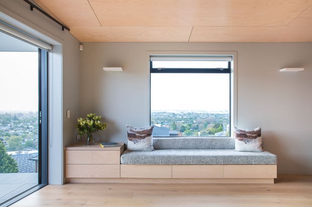 A bright, minimalist built-in window seat occupies the centre of a calm contemporary room. The upholstered bench includes integrated light timber drawers and cabinetry, with two cushions and a small plant on the adjacent low storage unit. Large windows provide an elevated suburban outlook, while pale timber flooring and a matching plywood ceiling create a warm, cohesive interior.