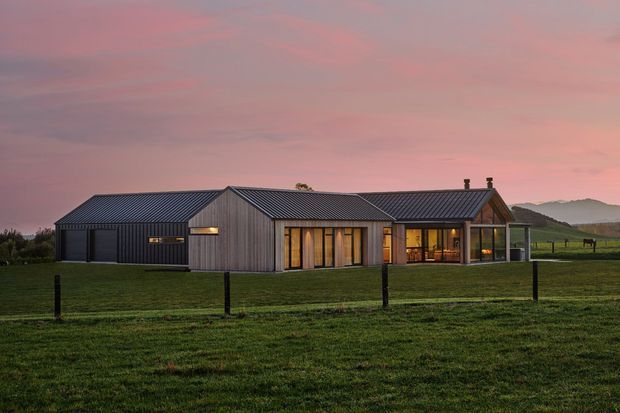 Single-storey contemporary rural residence viewed from across a fenced grassy field at dusk. The house has a low, elongated composition with multiple pitched gable roof volumes, dark vertical metal-like cladding, extensive warm-lit glazing, and a sheltered glazed living area. Rolling rural hills and open pasture surround the building.