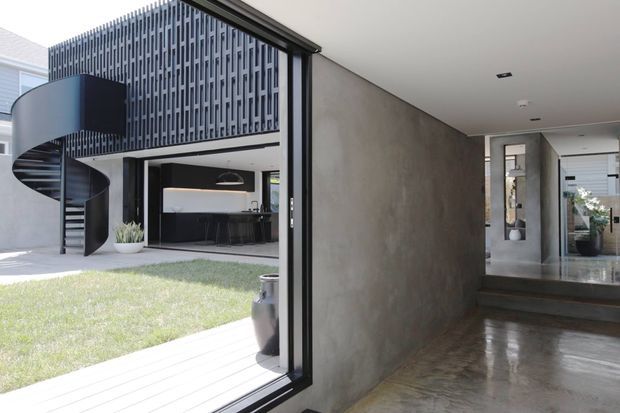 A contemporary residential entry and courtyard view featuring a polished concrete hallway, large black-framed glazing, a dramatic curved black spiral staircase, and a perforated vertical-screen facade. An open-plan kitchen and dining area is visible beyond the courtyard, with restrained neutral finishes and strong indoor-outdoor connection.