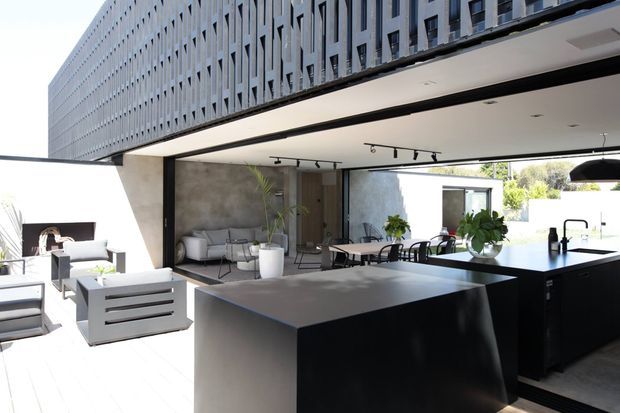 Contemporary open-plan indoor-outdoor living space with a dark minimalist kitchen island and black cabinetry opening onto a bright patio. The space features concrete-look walls, extensive black-framed glazing, track lighting, and a perforated upper facade creating a strong architectural overhang. Outdoor lounge and dining areas are visible beyond the kitchen.