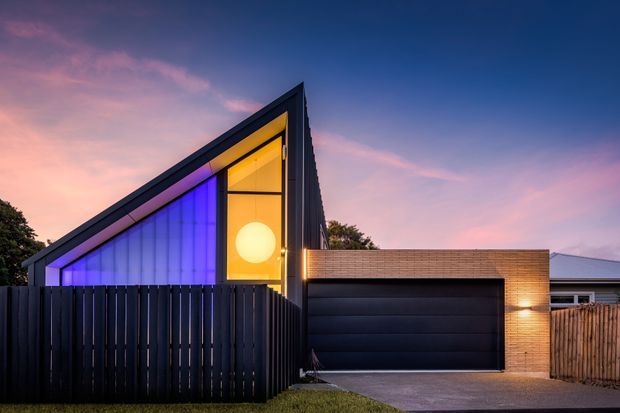 A contemporary single-storey residence viewed from the street at dusk. The sharply angled gable-like facade features extensive glazing illuminated in vivid blue and warm amber, contrasting with dark vertical fencing and a black double garage door. A warm-toned brick garage volume and timber side fencing complete the composition.