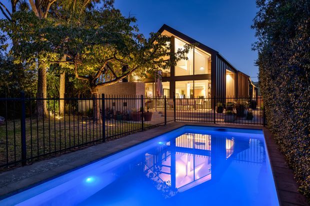 Twilight exterior view of a contemporary gabled residence beside a rectangular illuminated swimming pool. The home features dark vertical cladding, extensive floor-to-ceiling glazing, warm interior lighting, and a landscaped garden enclosed by a slim black metal fence.