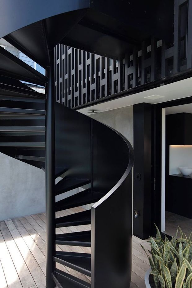 A dramatic contemporary interior courtyard or entry space featuring a sculptural black spiral staircase with open treads and a curved solid balustrade. Dark metal architectural elements contrast with pale concrete walls, timber-look decking, and a vertically patterned black-and-white upper-level screen. A black-framed opening reveals a minimalist dark bathroom or display niche, while indoor planting softens the composition.