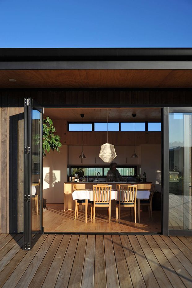 View from a timber deck through fully opened glazed doors into a warm, wood-lined dining and kitchen area. The room features a long dining table with timber chairs, a kitchen island, pendant lighting, cabinetry, and clerestory windows. The building has a strong indoor-outdoor connection, dark vertical timber cladding, broad roof overhangs, and a flat or very low-pitched roofline.