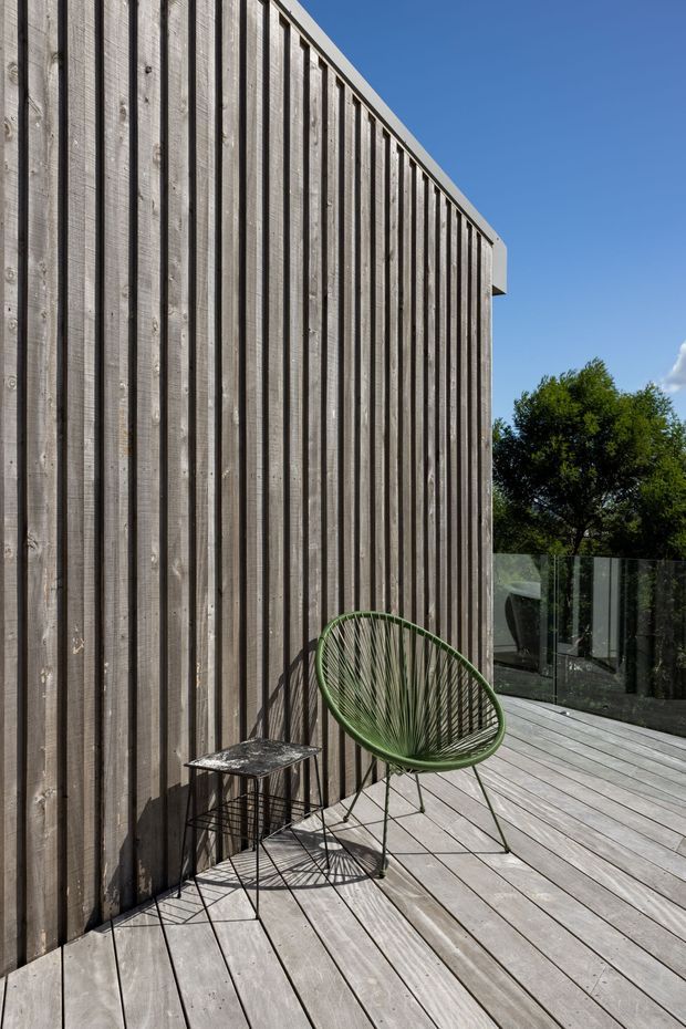 A sunlit exterior deck beside a tall facade clad in vertically oriented weathered timber boards. The deck has matching timber decking, a transparent glass balustrade, and a green woven outdoor lounge chair with a small dark metal side table. Trees and a clear blue sky are visible beyond the terrace.