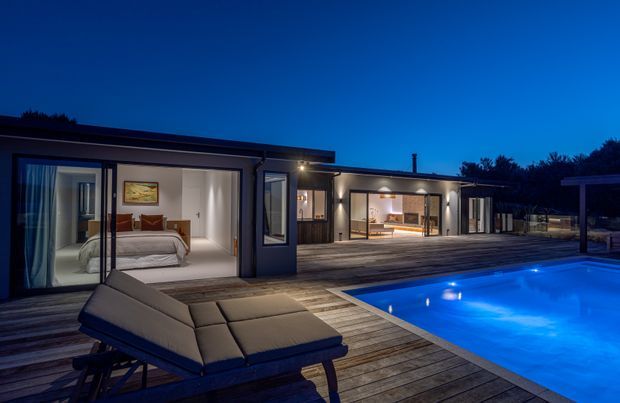 Twilight exterior view of a contemporary single-storey home opening onto a large timber deck and illuminated swimming pool. Floor-to-ceiling sliding glass doors reveal a bedroom on the left and an open living or dining area further along, while outdoor lounge furniture occupies the foreground. The home has a low-profile flat roof, dark framed glazing, and a strong indoor-outdoor connection.