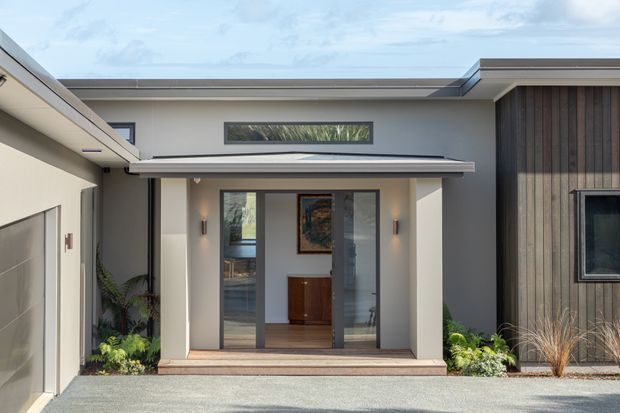 Contemporary single-storey residential entry facade with a covered entrance, glazed double doors, horizontal clerestory window, flat rooflines, light rendered walls, and a contrasting vertical timber-clad wall. Low-maintenance planting and gravel landscaping frame the entry.