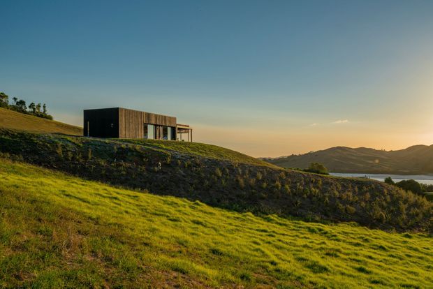A minimalist single-storey rectangular house set into a grassy, sloping rural landscape beside a body of water. The building has dark cladding, warm timber-clad sections, large glazed openings and a simple flat roof, photographed at golden hour with distant hills and shoreline beyond.