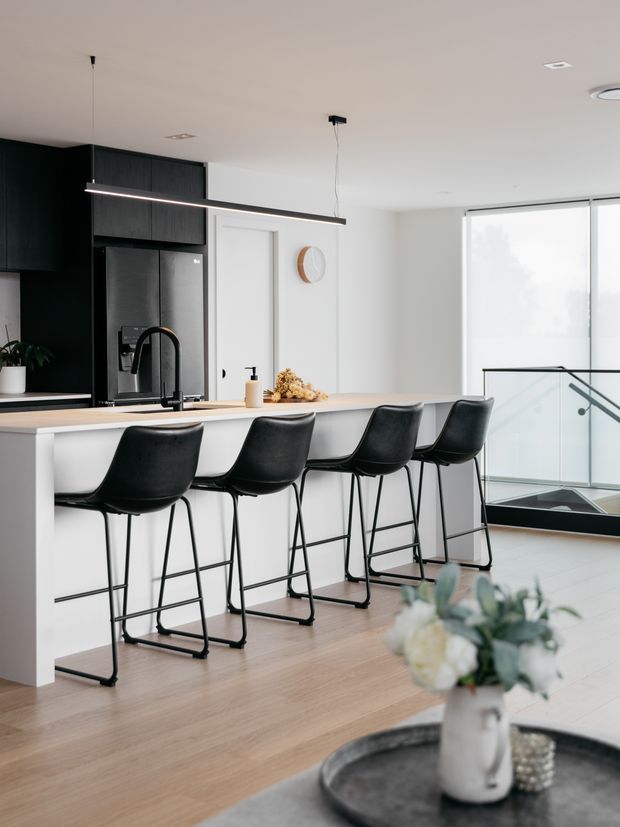 A bright, contemporary open-plan kitchen centered on a long white island with a pale stone benchtop and four black bar stools. The kitchen features matte black full-height cabinetry, a black refrigerator, dark integrated appliances, a black gooseneck tap, and a slim linear pendant light. Pale timber flooring, white walls, a round wall mirror, and a glazed stairwell with black framing complete the space. A floral arrangement on a foreground table is visible but secondary.