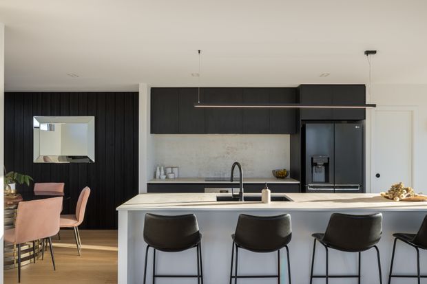 Contemporary open-plan kitchen and dining area with a large pale island bench, dark cabinetry, integrated black appliances, a textured light stone backsplash, and four black bar stools. A black vertical-panel feature wall with a geometric mirror sits behind the dining table, which has blush upholstered chairs. Minimal pendant lighting and warm timber flooring complete the space.