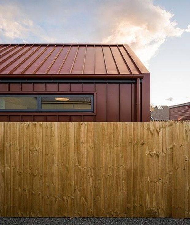 Exterior view of a compact building behind a tall timber privacy fence. The facade and pitched standing-seam metal roof are finished in a dark reddish-brown tone, with a long horizontal window in a dark frame and visible rainwater downpipe. Neighbouring rooflines appear in the background under a partly cloudy sky.