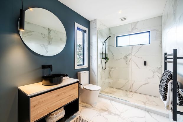 Contemporary bathroom with a floating timber vanity, black vessel basin and tapware, large round mirror, wall-hung toilet, and a walk-in shower. White marble-look wall and floor tiles contrast with a dark blue painted feature wall and black towel ladder.