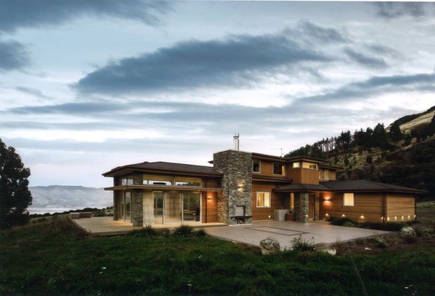 Exterior view of a contemporary multi-level hillside house with broad low-pitched roof planes, extensive floor-to-ceiling glazing, warm timber cladding, stone feature walls, and an integrated double garage. The home sits on a sloped landscaped site with a distant mountainous or coastal landscape beyond.