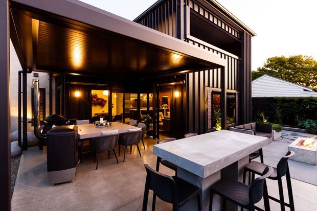 A sophisticated covered outdoor entertaining area adjoining a contemporary multi-level home. The space features dark vertical cladding, timber-lined soffits, warm integrated lighting, an outdoor dining setting, a large stone-look dining table, barbecue cabinetry, and a built-in fire feature on a paved terrace.