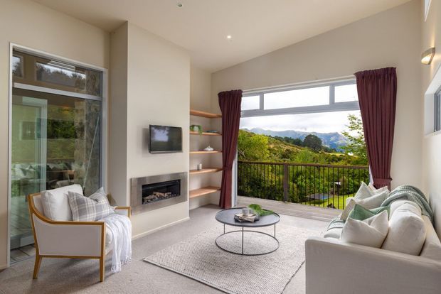 Bright contemporary living room with white upholstered seating, a round glass coffee table, pale carpet, built-in shelving, wall-mounted television and a recessed fireplace. Full-height glazing and an open balcony provide views toward a lush, mountainous landscape, while burgundy curtains add contrast.