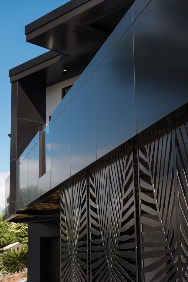 A close-up exterior view of a contemporary building featuring dark reflective cladding, sharp cantilevered roof forms, and a decorative metal privacy screen with an organic leaf-like pattern. Strong geometric lines, black and charcoal finishes, and reflective glazing dominate the facade against a clear blue sky.
