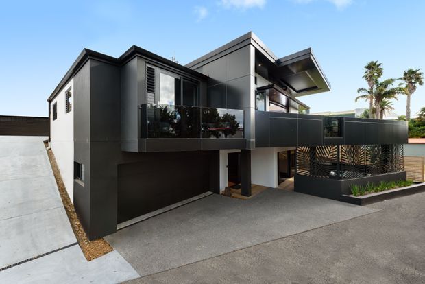 Exterior view of a contemporary multi-level residence with dark charcoal cladding, projecting box-like forms, angled roof planes, upper-level balconies with dark privacy screens, and an integrated ground-level garage. The home fronts a paved driveway and includes tropical palms visible in the background.