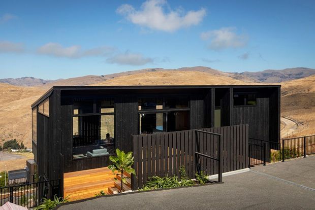 A compact contemporary dark-clad house set on a steep, elevated site, with large black-framed windows, a timber deck and entry steps, screened outdoor areas, and surrounding dry mountainous terrain.