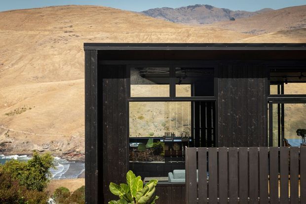 Exterior view of a dark, timber-clad modern structure set against a dry mountainous landscape. Large black-framed glazed openings reveal a partially visible interior, while a dark timber privacy screen and planting occupy the foreground. A rocky river or stream is visible below the building.