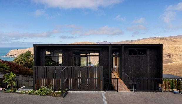 Exterior view of a compact modern single-storey residence with dark vertical timber cladding, a flat roof, large glazed openings, and a timber privacy fence and deck. The home is set on a coastal, elevated site with ocean and dry hills visible beyond.
