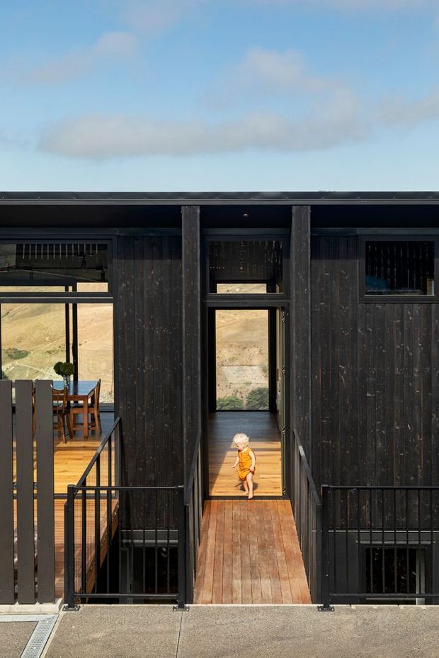 Exterior view of a dark timber-clad, flat-roofed multi-level building with a central elevated timber walkway and black metal balustrades. Large windows reveal a dining area and warm timber flooring inside, while a child stands in the central doorway. The building is set against a bright open landscape and sky.