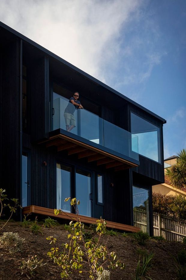 Exterior view of a contemporary two-storey house on a sloping site, featuring dark vertical cladding, extensive glazing, a projecting upper-level balcony with glass balustrade, exposed timber beams beneath the balcony, and a simple low-pitched roofline. Native-style planting and fencing surround the building.