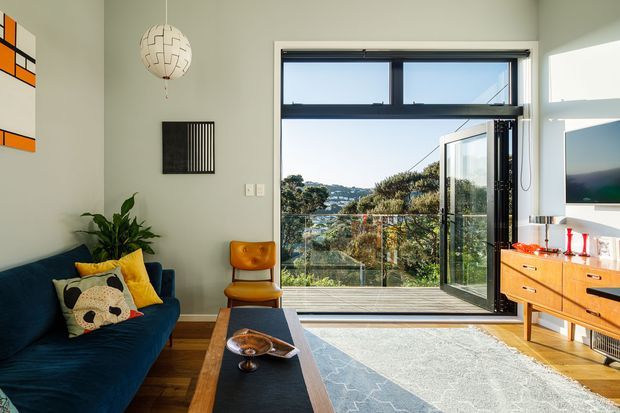 A bright, compact mid-century-inspired living room with a deep blue sofa, timber flooring, a patterned rug, and warm timber furniture. The room opens through large black-framed folding doors to a balcony or terrace with a leafy outlook. A wall-mounted television, pendant light, artwork, indoor plant, and decorative furnishings complete the space.