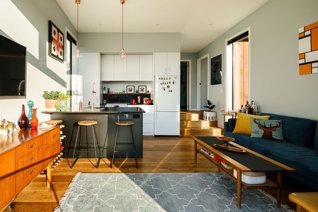 A bright open-plan living and kitchen area with a mid-century-inspired interior. The space features warm timber flooring, a white galley kitchen with a dark island and bar stools, a teal sofa with colourful cushions, a low timber coffee table, a timber sideboard, patterned rug, and large windows bringing in abundant natural light. Abstract artwork, pendant lights, and indoor plants add character.