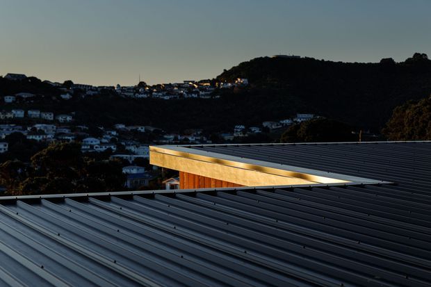 Exterior architectural view focused on overlapping low-pitched metal roofs with pronounced linear ribbing. A warm timber-clad upper volume and glazed or open edge are visible between the roof planes, with a densely settled, hilly suburban landscape in the background at dusk.