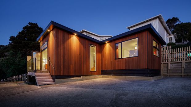 A contemporary single-storey residential extension or house viewed from the driveway at dusk. The compact angular form features warm vertical timber cladding, dark trim and base detailing, large windows, exterior wall lighting, and a raised entry deck with steps. A taller light-coloured upper roof volume is visible behind the main timber-clad structure, with landscaped trees and fencing surrounding the site.