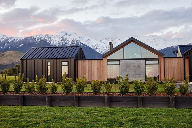 A low-profile contemporary residential building with several connected gabled volumes, photographed from the landscaped front yard. The facade combines dark vertical cladding, warm timber boards, large glazed gable sections, and dark standing-seam metal roofs. Formal hedges and raised garden beds line the foreground, with snow-covered mountains forming the distant backdrop.