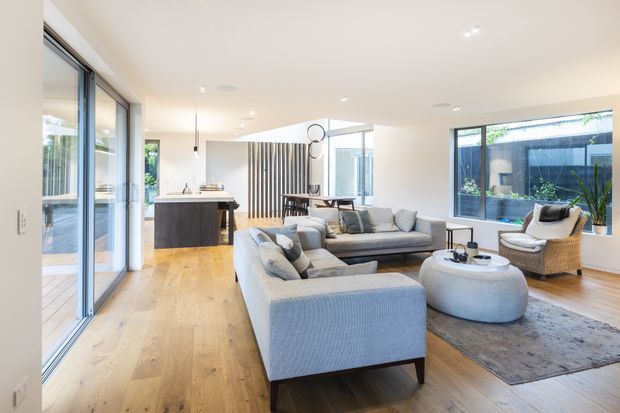 Bright open-plan contemporary interior featuring a spacious living room, kitchen and dining area. The room has pale timber flooring, white walls and ceiling, extensive floor-to-ceiling glazing, a large light-grey sectional sofa, rounded ottoman coffee table, woven lounge chair, and a dark area rug. A compact island kitchen with dark cabinetry and a pale benchtop sits behind the living area, with vertical timber screening near the dining space. The space opens to a planted outdoor courtyard visible through the windows.