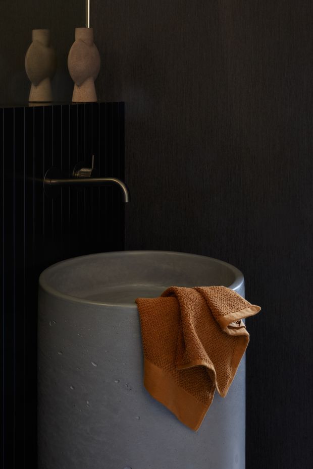 Moody close-up of a contemporary bathroom featuring a freestanding cylindrical concrete basin, matte black vertically grooved wall and splashback surfaces, a dark metal wall-mounted tap, and warm terracotta towels. Two sculptural ceramic vessels sit on the ledge above the basin.