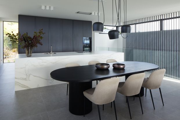 Contemporary open-plan kitchen and dining area with a large white veined stone island, dark charcoal cabinetry, a black sculptural oval dining table, and four upholstered dining chairs. Multiple black pendant lights hang above the table, while full-height vertical glazing or screening lines the right side of the room. The space has a refined monochromatic palette and abundant natural light.