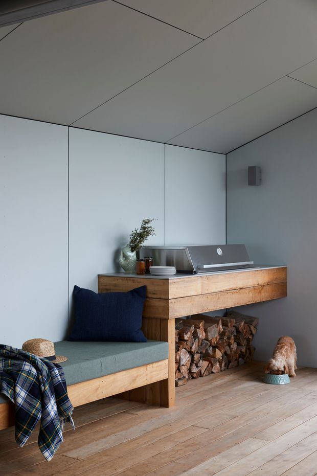A compact, softly lit interior corner with pale grey paneled walls and ceiling, wide light timber floorboards, and a built-in timber bench with a muted green cushion. A dark navy cushion and checked throw add contrast. Above the bench is a substantial timber console or hearth-like joinery unit with an open compartment stacked with firewood and a sleek grey metal appliance or storage element on top. Minimal decorative objects and a small dog create a calm, warm, refined atmosphere.