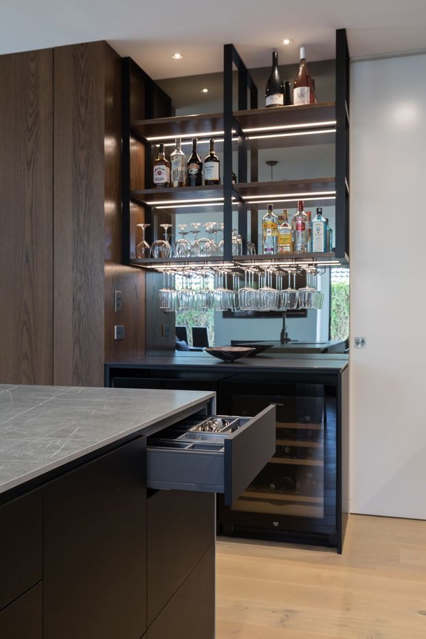 A refined residential bar area integrated into a dark timber and black cabinetry kitchen. The space features open black metal shelving with illuminated timber shelves, liquor bottles, suspended wine and cocktail glasses, mirrored backing, a wine fridge, and a pull-out drawer with bar accessories. A large stone-look island and pale timber flooring are visible in the foreground.
