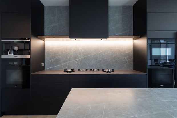 A dark, sophisticated contemporary kitchen with full-height black cabinetry, integrated appliances, a four-burner gas cooktop, and a large central island. The backsplash and island surface feature veined grey stone, while concealed linear lighting illuminates the cooking zone and open shelving displays glassware.