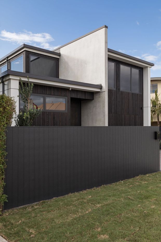 Exterior photograph of a contemporary two-storey townhouse or compact residence. The facade combines dark vertical timber-look cladding, smooth light rendered walls, large dark-framed windows, and a tall asymmetrical parapet wall. A dark privacy fence and small landscaped planting sit in front of the building.