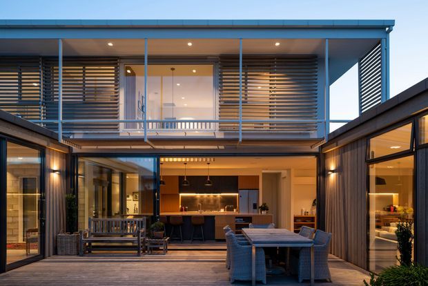 A contemporary two-storey courtyard home viewed from an outdoor entertaining deck. The central open-plan area contains a kitchen with an island and pendant lights, a dining table, and glazed living spaces, while the upper level features extensive glazing, a balcony, and horizontal timber screening. Dark framed glazing and timber decking create a warm, modern indoor-outdoor setting.