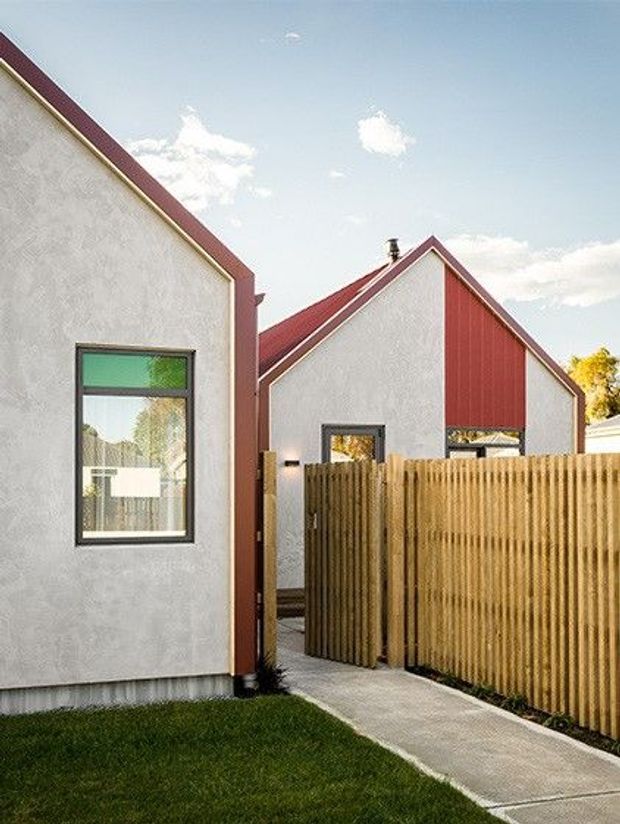 Exterior view of a contemporary residential building composed of compact gabled volumes. The facades are light grey textured render with contrasting red vertical metal cladding and red roof-edge trims. A timber privacy fence and gate line a narrow concrete-paved walkway, with a small lawn in the foreground. The image is photographed in bright daylight and emphasizes the building envelope and pedestrian entry.